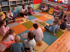 Przedszkolaki z grupy Promyczki siedzą na dywanie i słuchają bajek czytanych przez nauczyciela.