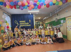Przedszkolaki z grupy: Zuchy, Krasnale i Promyczki wraz z Ciociami oraz pedagogiem specjalnym stoją na korytarzu przedszkolnym prezentują żółte stroje.