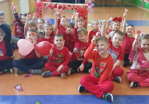 Grupa Zuchy - zdjęcie grupowe przedszkolaków siedzących siadem skrzyżnym w czerwonych strojach a w dłoniach trzymają serduszka.
