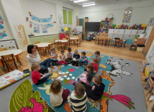Przedszkolaki z grupy Kamyczki siedzą na dywanie wraz z pedagogiem specjalnym i aktywnie uczestniczą w zajęciach.