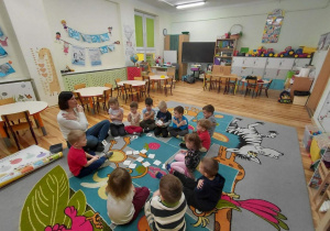 Przedszkolaki z grupy Kamyczki siedzą na dywanie wraz z pedagogiem specjalnym i aktywnie uczestniczą w zajęciach.