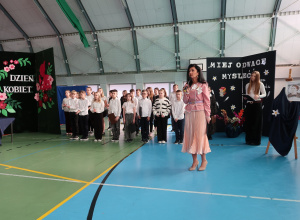 Pani Dyrektor przemawia do zgromadzonej publiczności stojąc na środku z tyłu są obecni uczniowie klasy IV.