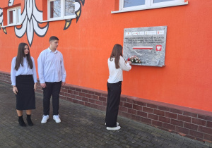 Zastępca przewodniczącej przed budynkiem szkoły wraz z delegacją uczniów składa kwiaty pod tablicą upamiętniającą uczniów, rodziców i nauczycieli walczących o szkołę polską w gminie Rzeczyca.