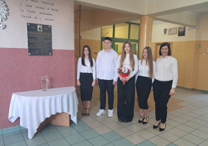 Delegacja uczniów klasy VIII a i VIII b wraz z Opiekunem Samorządu oraz Przewodniczącą która trzyma kwiaty, na korytarzu szkolnym obok tablicy Narcyzy Żmichowskiej.