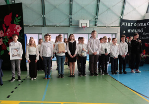 Uczniowie stojąc na środku prezentują zdjęcie Narcyzy Żmichowskiej oraz symbole poetki tj. książka, pióro, światło i kwiatek.