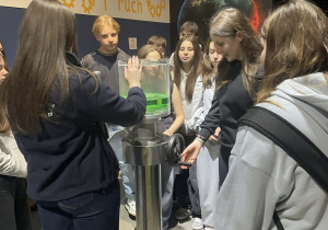 Grupa uczniów z zaciekawieniem patrzy na instruktaż edukatorki Experymentarium.