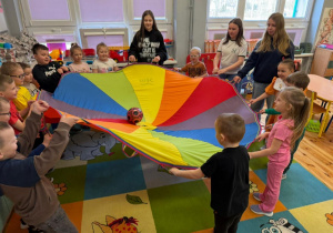 Uczennice klasy VI i VII a z przedszkolakami z grupy Promyczki bawią się chustą klanza w środku chusty znajduje się piłka.