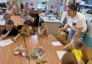 Pani Klaudia udziela dzieciom siedzącym przy stolikach wskazówki dotyczące warsztatów.
