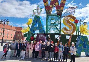 Uczniowie klasy pierwszej ze Szkoły Filialnej w Sadykierzu z wychowawcą stoją przed dużym napisem Warszawa.