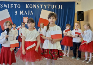 Uczennice klasy pierwszej trzymają w ręku flagę i recytują wiersze podczas uroczystości.