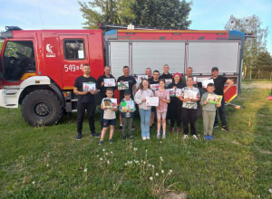 Uśmiechnięci, zadowoleni strażacy trzymający otrzymane prace na tle wozu strażackiego.