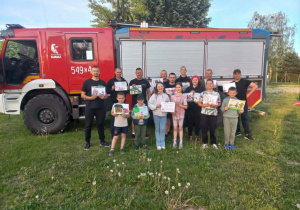 Uśmiechnięci, zadowoleni strażacy trzymający otrzymane prace na tle wozu strażackiego.