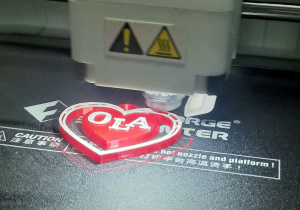 Drukarka 3D drukuje czerwone serce z białym napisem nakładając kolejne warstwy filamentu.